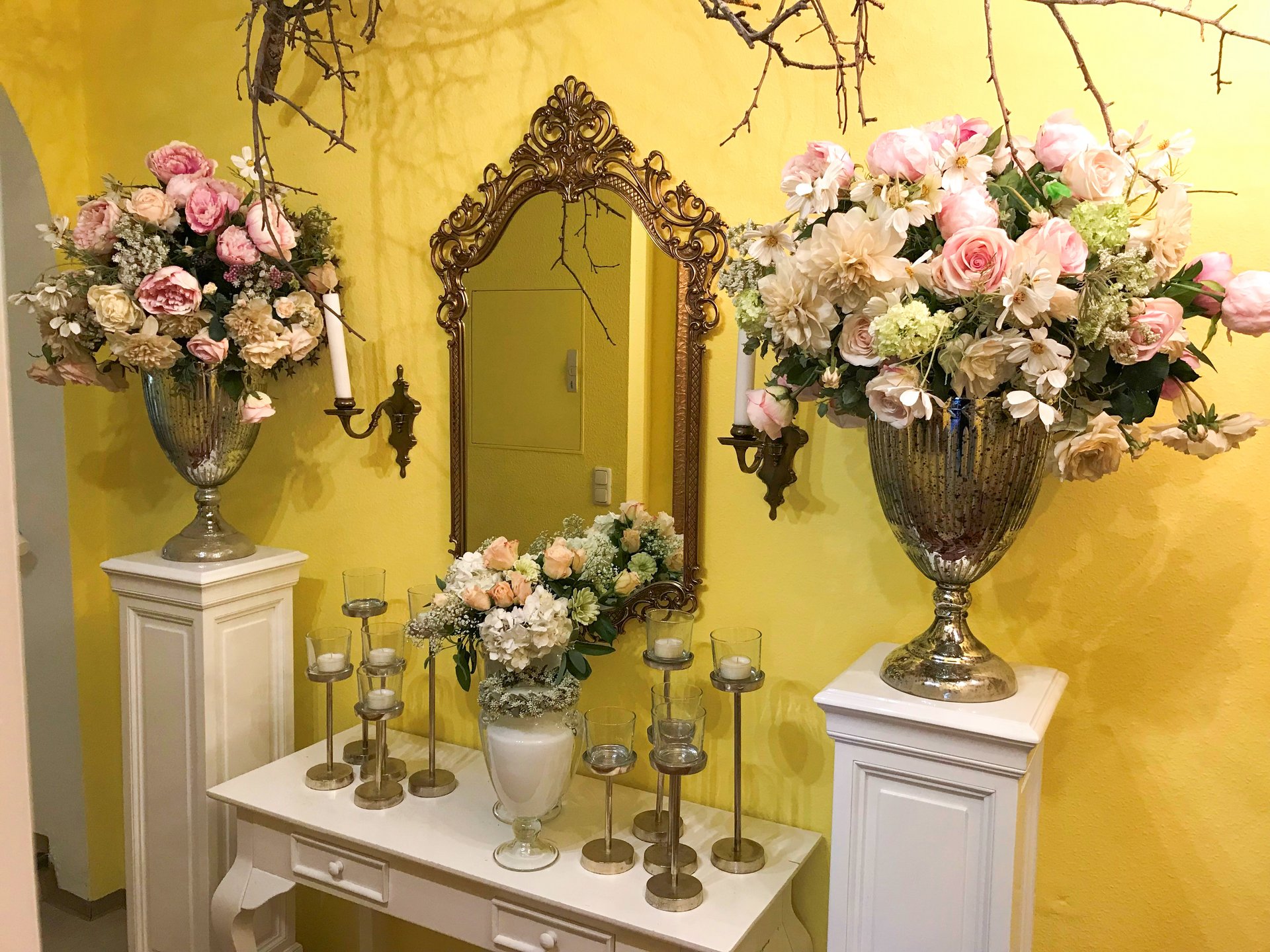 Aufnahme eines weißen Schminktisches, der vor einer gelben Wand steht. Links und rechts neben dem Tisch stehen zwei Kommoden. Tisch und Kommoden sind mit Blumen und Kerzen dekoriert. An der Wand hängt ein goldener Spiegel.